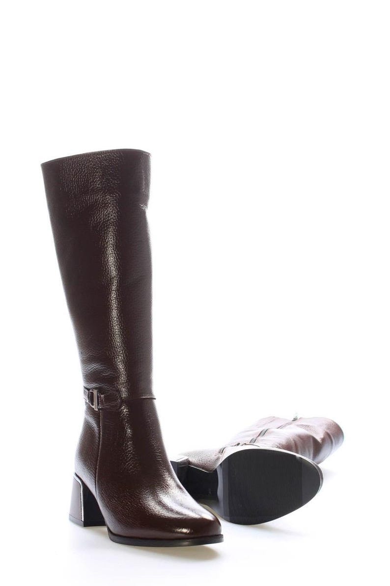 May include: A pair of knee-high brown leather boots with a glossy finish. One boot stands upright, while the other lies on its side. Each boot features a silver buckle and a block heel.