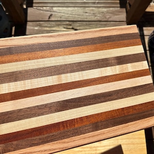 Può includere: Un tagliere rettangolare in legno con strisce alternate di legno chiaro e scuro. Il tagliere è fatto di diversi tipi di legno, tra cui acero, noce e ciliegio.