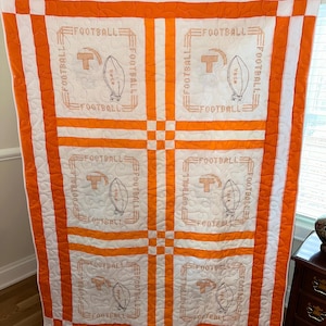 May include: A white and orange patchwork quilt with six embroidered squares featuring the words "FOOTBALL" and a stylized football and letter "T".