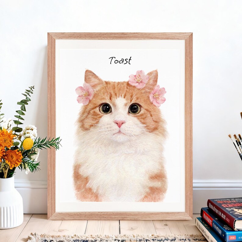 Puede incluir: Una impresi&oacute;n enmarcada de acuarela de un gato con pelaje naranja y blanco, con una corona de flores rosas. La palabra "Toast" est&aacute; escrita encima de la cabeza del gato. El marco es de color marr&oacute;n claro.