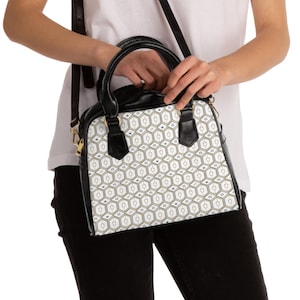Puede incluir: Bolso con estampado en blanco y negro con asa superior y bandolera. El bolso presenta un diseño geométrico en dorado y negro, con fondo blanco. El bolso lo sostiene una persona que lleva una camisa blanca y pantalones negros.
