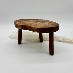 May include: A rustic wooden stool with a natural wood finish and three legs. The stool has an oval-shaped top and is made of solid wood.