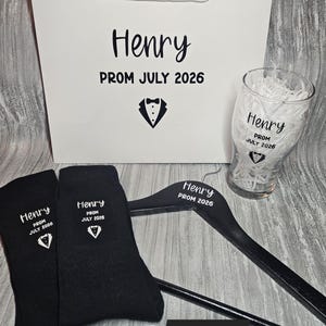 May include: A prom gift set featuring a white bag, pint glass, black hanger, and black socks, all personalised with "Henry PROM JULY 2026" and a tuxedo graphic. The set is labelled "Set 2".