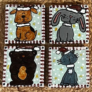May include: Set of four square ceramic coasters featuring cartoon animals in Santa hats. The dog, bunny, bear, and cat are painted on a light blue background with white snowflakes and yellow stars. Each coaster has a white border with a red and brown dotted pattern.