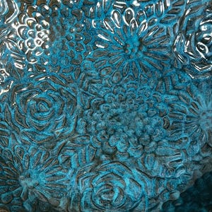 May include: Close-up of a teal ceramic bowl with a floral pattern. The bowl's surface features embossed flowers and leaves in varying sizes, with a glossy finish. The rim of the bowl is a natural woven material.