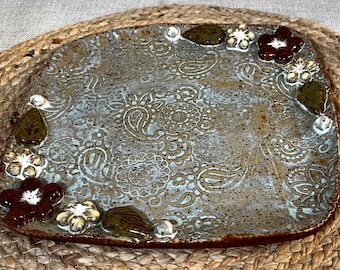 May include: Rectangular ceramic serving platter with a paisley pattern in shades of brown and blue. The edges are decorated with brown and white flowers, green leaves, and small white accents. The platter is placed on a woven mat.