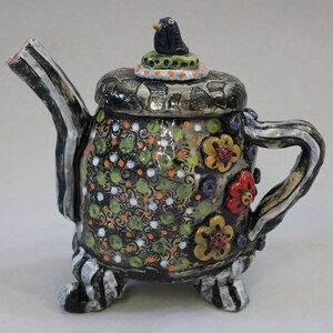 May include: A decorative ceramic teapot with a black, white, and grey striped spout and handle. The body is adorned with green, orange, and white floral patterns and flowers. A small bird figurine sits atop the lid. The teapot is supported by three small feet.