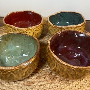 May include: Four handmade ceramic bowls with organic shapes and textured exteriors. Each bowl has a unique interior color: red, green, teal, and purple. The bowls are arranged on a woven placemat, showcasing their rustic charm.