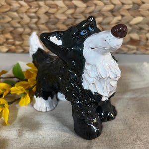 May include: A ceramic dog figurine with a black and white coat, blue eyes, and a brown nose. The dog is standing, with its head tilted slightly upwards. The figurine has a glossy finish and is set against a neutral background.