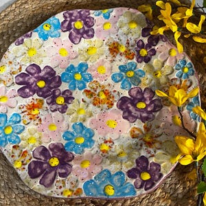 May include: A ceramic plate with a floral design. The plate features a variety of colorful flowers in shades of purple, blue, yellow, and pink. The flowers are arranged on a white background with speckled accents. The plate has an irregular shape.