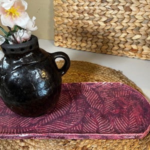 May include: A black ceramic vase with two handles, holding white and pink flowers, sits on a rectangular, burgundy tray with a leaf pattern. The tray rests on a woven mat, with a wicker basket in the background.