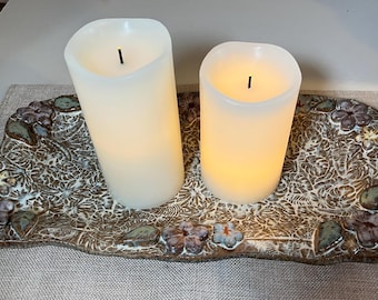May include: Two white pillar candles with black wicks are sitting on a decorative ceramic tray with a floral design. The tray is brown with white and light brown accents.