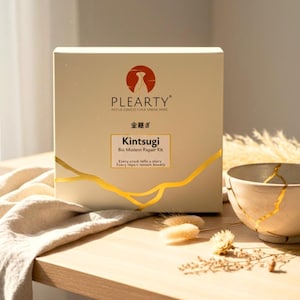 May include: A Kintsugi Bio Modern Repair Kit box with gold accents and the text "PLEARTY" and "Kintsugi". A ceramic bowl with gold-filled cracks sits nearby. The box and bowl are on a wooden surface.