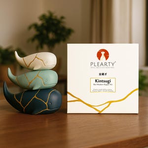 May include: Three whale figurines in cream, teal, and navy blue, stacked on a wooden surface. Each whale has gold-colored crack patterns. A white box with the text "PLEARTY Kintsugi Bio Modern Repair Kit" is to the right.