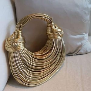 Könnte beinhalten: Eine goldfarbene Handtasche mit einem einzigartigen Design. Die Tasche hat einen abgerundeten Griff und einen Körper, der aus zahlreichen parallelen, vertikalen Strängen besteht. Der Griff ist oben verknotet, was ein dekoratives Element darstellt.