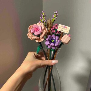 May include: A clear glass vase holding a bouquet of colorful flowers. The flowers are made of plastic and include pink roses, purple and pink tulips, and lavender sprigs. A small card with text is also included.