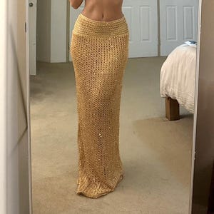 May include: A floor-length, gold-colored skirt with a knitted design. The skirt has a wide waistband and a straight silhouette. The fabric appears to have a subtle shimmer, catching the light. The skirt is made of a soft, flowing material.
