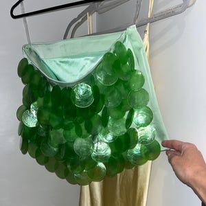 May include: A mint green handbag with a shoulder strap, decorated with iridescent green circular sequins. The bag is hanging on a hanger against a white wall. The bag's design is eye-catching and unique.