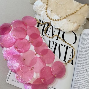 May include: A close-up of two decorative accessories. One is a white, scalloped clutch with a gold chain. The other is a pink, sequined accessory. Both are on top of a magazine with the text "PONTO DE VISTA".