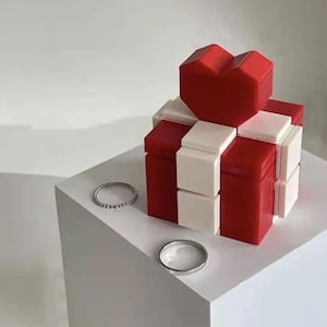 May include: A red, white, and silver gift box with a heart-shaped top. The box is made of red and white blocks. Two silver rings and a silver bracelet are on the white surface.