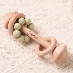 以下が含まれることがあります： 「Emma」の名前が刻まれた木製のベビーラトル。明るい木目調で、緑色のビーズと円形の持ち手が付いています。乳幼児向けに設計されています。