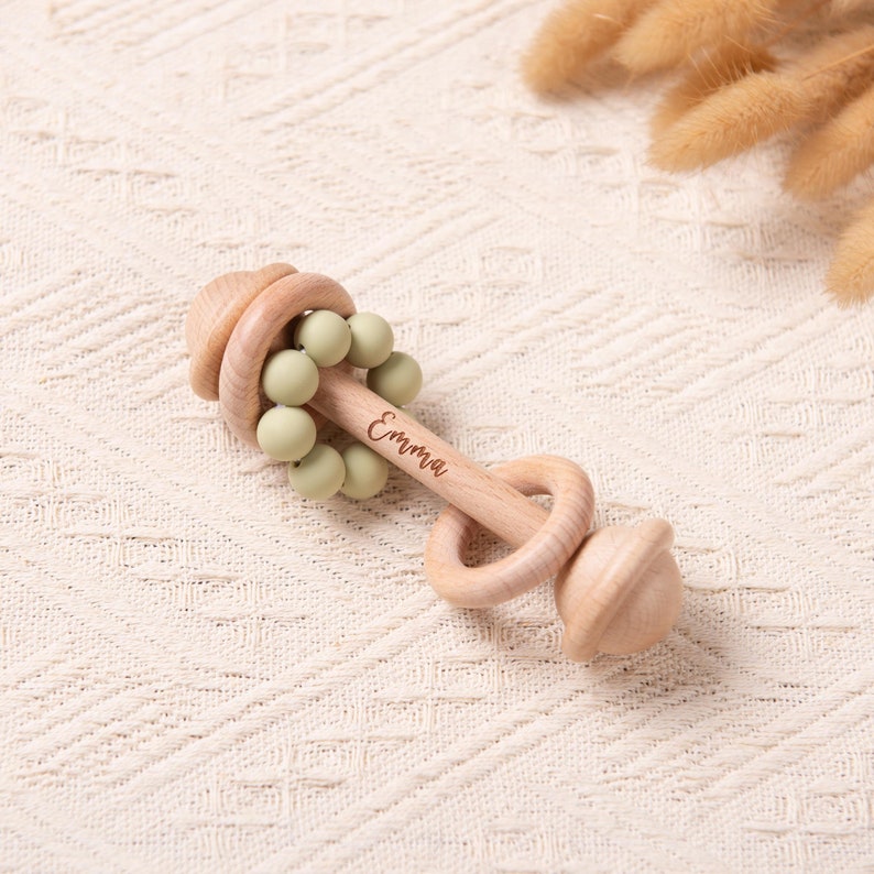 May include: Wooden baby rattle with the name "Emma" engraved on the handle. The rattle features a light wood finish, a series of green beads, and a circular handle. The rattle is designed for infants and toddlers.