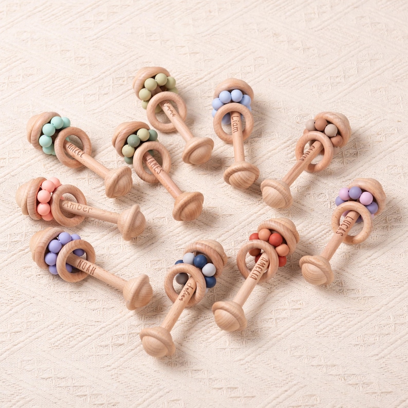 May include: A collection of wooden baby rattles with colorful silicone beads. Each rattle has a wooden handle and a ring with a name engraved on it. The beads come in various colors, including pink, blue, and green.