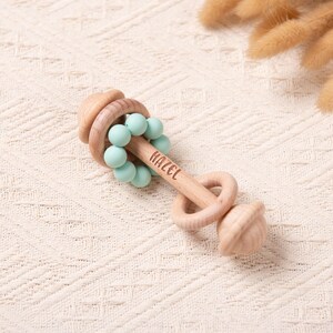 May include: A wooden baby rattle with a light wood finish. The rattle features a series of light blue beads and the name "HAZEL" engraved on the handle. The design includes a ring and an acorn-shaped end.