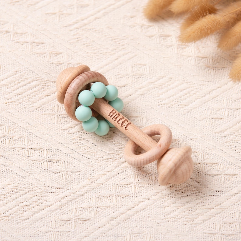 May include: A wooden baby rattle with a light wood finish. The rattle features a series of light blue beads and the name "HAZEL" engraved on the handle. The design includes a ring and an acorn-shaped end.