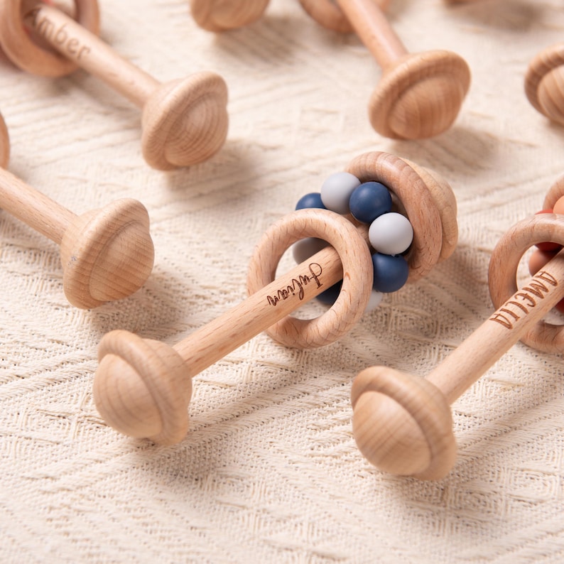 May include: Wooden baby rattles with a natural finish. Each rattle features a handle, a round head, and a ring with beads. Some rattles have names engraved on them, such as "Julian" and "Amelia". The beads are in shades of blue and white.