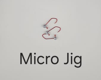 Może przedstawiać: Trzy czerwone haczyki do wędkowania micro jig z srebrnymi główkami jig są pokazane na białej powierzchni. Tekst "Micro Jig" jest wydrukowany na czarno poniżej.