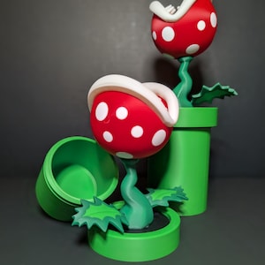May include: Two 3D printed red and white Piranha Plants from the Super Mario Bros. video game series. The plants are in green pipes with green leaves.