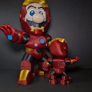 May include: Two red and gold figurines of Mario in an Iron Man suit. The larger figure has a blue circle on its chest and a yellow helmet. The smaller figure is in a similar pose. The background is dark.