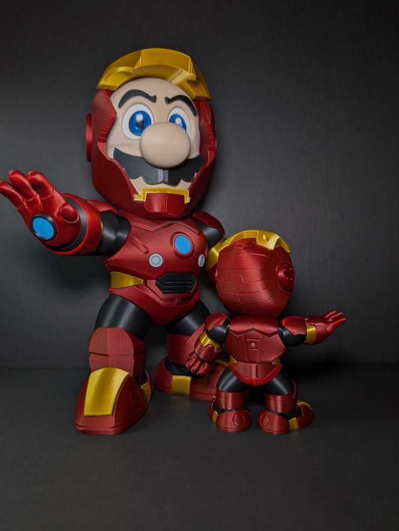 May include: Two red and gold figurines of Mario in an Iron Man suit. The larger figure has a blue circle on its chest and a yellow helmet. The smaller figure is in a similar pose. The background is dark.