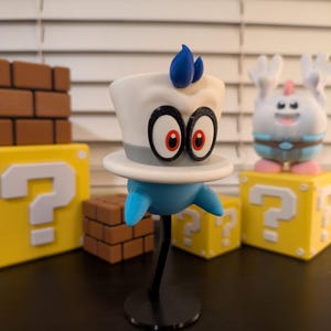 May include: A 3D printed figurine of a character from the Super Mario video game series. The character is wearing a white hat with a blue flame on top and has red eyes. The figurine is standing on a black stand.