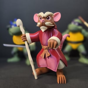 May include: A detailed figurine of a rat character in a red robe with white trim, holding a wooden staff. The rat has brown fur, pink ears, and a white beard. Two other figures are blurred in the background.