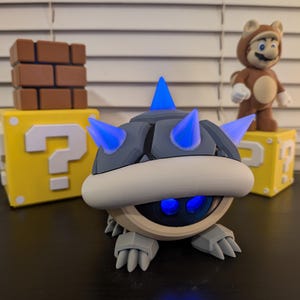 May include: A blue and gray 3D printed figurine of a Koopa Troopa from the Super Mario Bros. video game series. The figurine has blue spikes and glowing blue eyes. The figurine is sitting on a black surface.