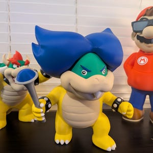 May include: Three yellow and green Bowser figures from the Super Mario Bros. video game franchise. The figure on the left is holding a blue and silver scepter. The figure in the center has blue hair and is holding a blue and silver scepter. The figure on the right is wearing a red shirt with a white M logo and blue pants.