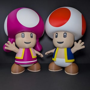 May include: Two figurines of Mushroom Kingdom characters. One has a pink mushroom cap with a white spot, a pink dress, and brown shoes. The other has a red and white mushroom cap, a blue vest, and brown shoes. Both have smiling faces.