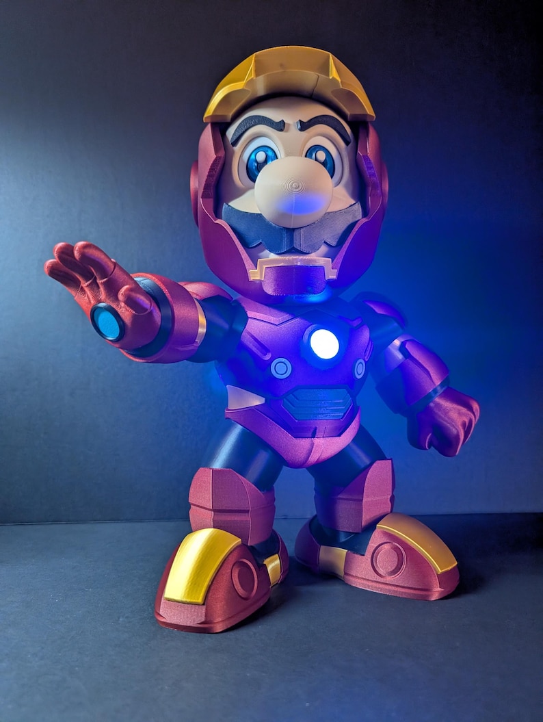 May include: A 3D-printed figurine of Mario in an Iron Man suit. The figure is primarily red and gold, with blue accents and glowing lights. The helmet and boots are gold, while the body is red. The figure is posed with one arm outstretched.