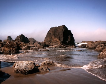 May include: A rocky coastline with large, dark grey rocks jutting out of the water. The water is a light blue-grey colour and there are white waves crashing against the rocks. The sky is a clear blue.