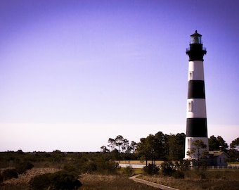 May include: A black and white striped lighthouse stands tall against a clear blue sky. The lighthouse is surrounded by green trees and grass.