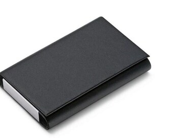 Puede incluir: Un porta tarjetas de cuero negro con un borde de metal plateado. El porta tarjetas está cerrado y se encuentra sobre una superficie blanca.