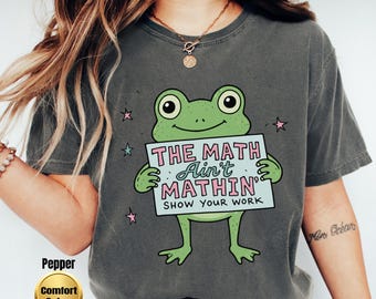 May include: A charcoal gray t-shirt featuring a green frog illustration holding a light blue sign that reads THE MATH Ain't MATHIN' SHOW YOUR WORK in pink and black lettering, accented by small pink and teal stars surrounding the frog graphic.