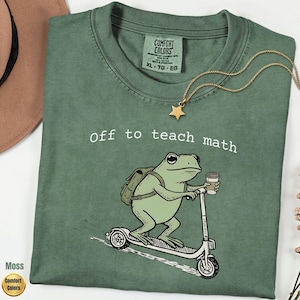 May include: A sage green t-shirt featuring a graphic of a frog wearing a backpack and riding a scooter while holding a coffee cup. The text above the frog reads "off to teach math" in white letters. The shirt has a Comfort Colors brand label.