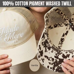 May include: A vanilla-colored trucker hat made of 100% cotton pigment washed twill. The front features white embroidered text reading Made for More Ephesians 2:10. The hat has a mesh back and an interior lining with a brown and white cow print pattern.