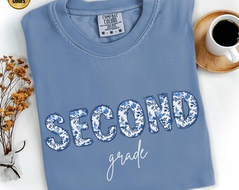 Könnte beinhalten: Ein blaues Comfort Colors T-Shirt mit der Aufschrift "SECOND grade" auf der Vorderseite. Das Wort "SECOND" ist in großen Buchstaben mit floralem Muster gehalten, und "grade" ist in einer Schreibschrift. Eine Tasse Kaffee steht auf einem Holzuntersetzer.
