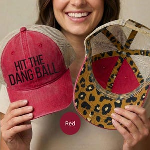 May include: Two red baseball caps with mesh backs are displayed. One cap has the text "HIT THE DANG BALL" in black. The other cap has a leopard print design on the underside. The caps are red.