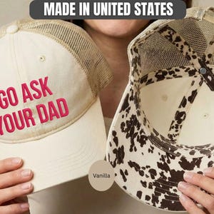 K&ouml;nnte beinhalten: Zwei vanillefarbene Trucker-H&uuml;te. Auf einem Hut steht der Text "GO ASK YOUR DAD" in rosa Stickerei. Der andere Hut hat ein Kuhmuster auf der Innenseite. Beide H&uuml;te haben einen Mesh-R&uuml;cken und einen gebogenen Schirm. Hergestellt in den Vereinigten Staaten.