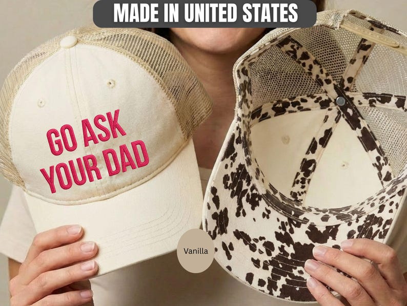 K&ouml;nnte beinhalten: Zwei vanillefarbene Trucker-H&uuml;te. Auf einem Hut steht der Text "GO ASK YOUR DAD" in rosa Stickerei. Der andere Hut hat ein Kuhmuster auf der Innenseite. Beide H&uuml;te haben einen Mesh-R&uuml;cken und einen gebogenen Schirm. Hergestellt in den Vereinigten Staaten.